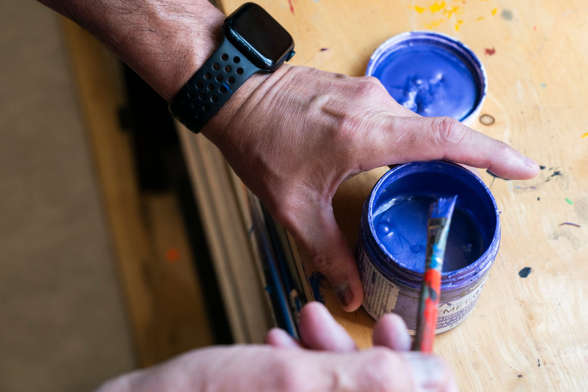 A photo of a man's hand holding a cup with blue paint. His other hand is seen holding a paintbrush. 