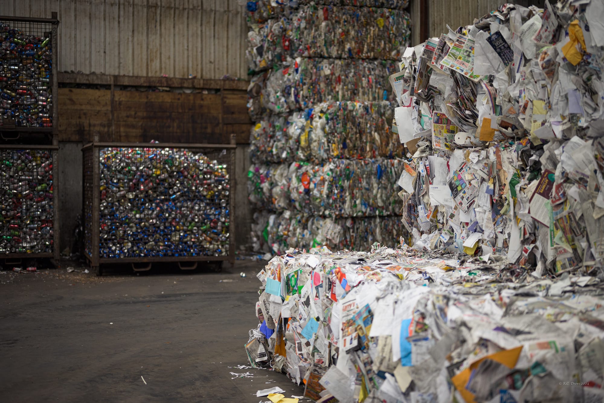 Bales of paper and bins and bales of aluminum cans are stacked along a recycling facility's wall.