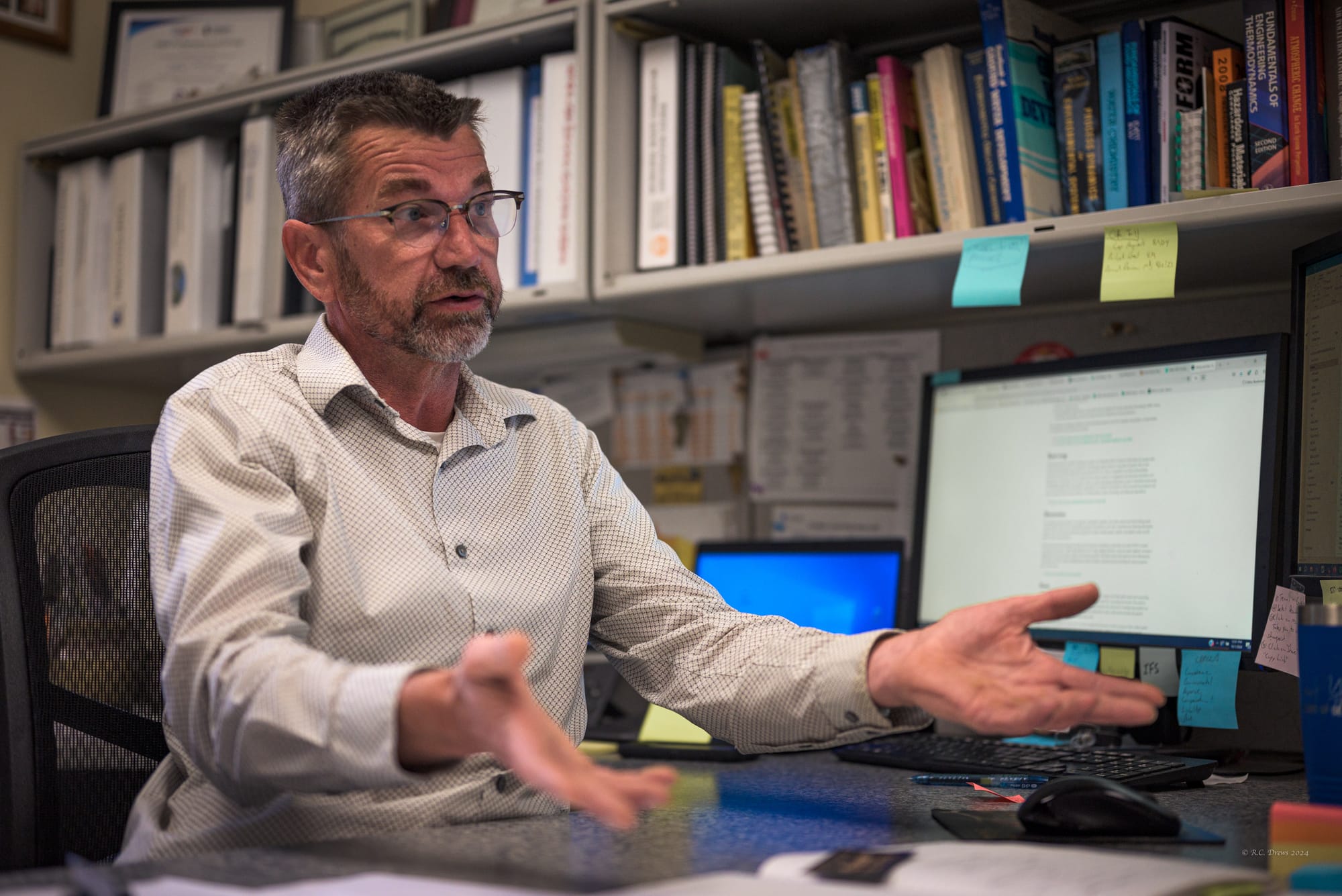 Man with glasses and a beard talks while seated behind a desk. 