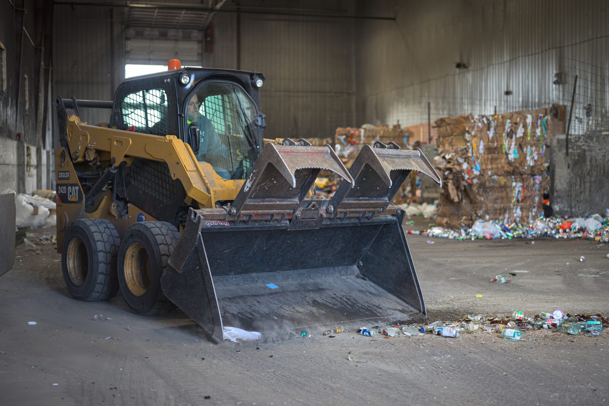 A worker drives a skid steer to push glass containers into a bunker at a recycling facility.