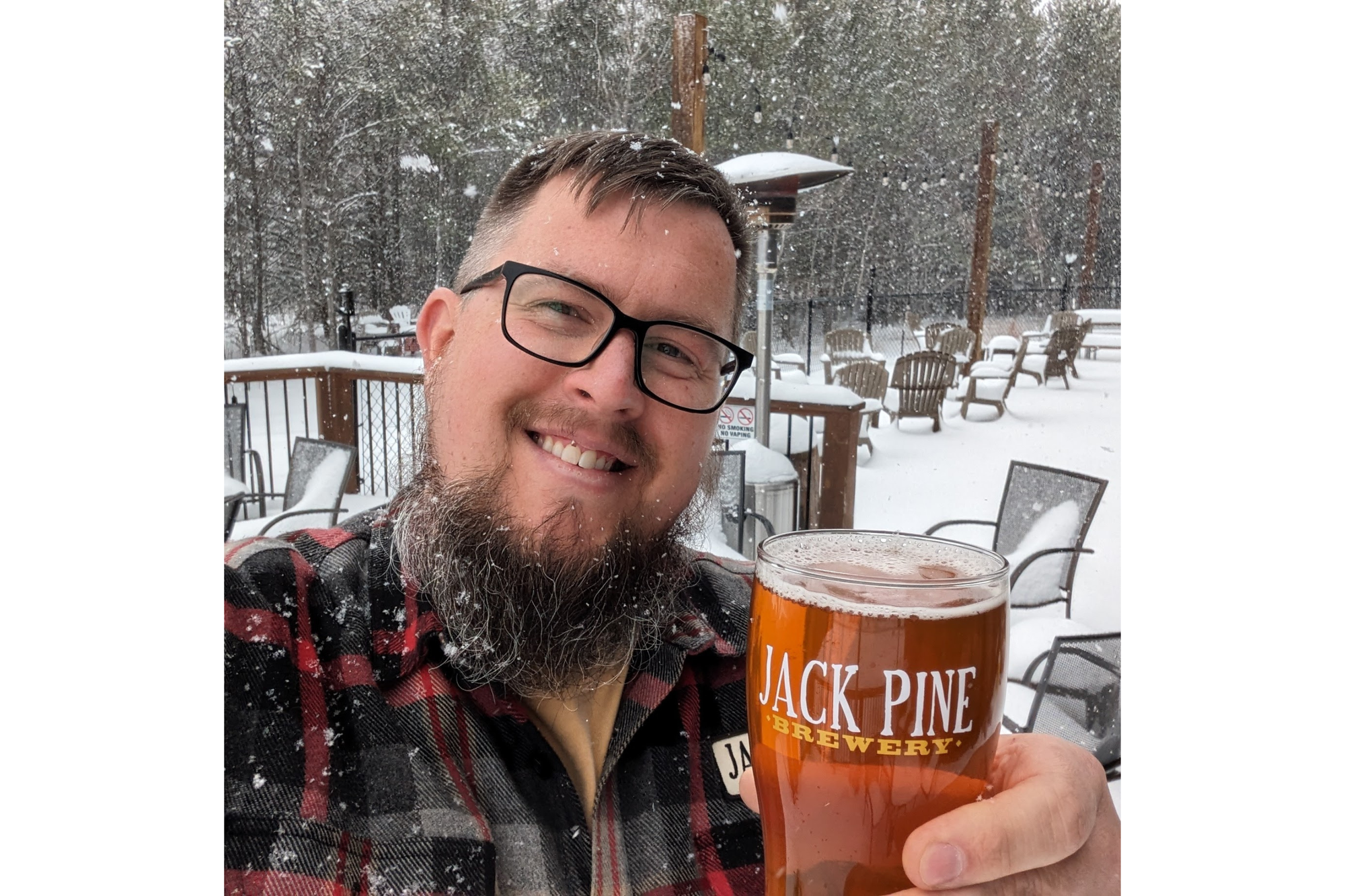 Man with a beard and glasses smiles for a photo. He's holding a glass of beer in a snowy backdrop. 