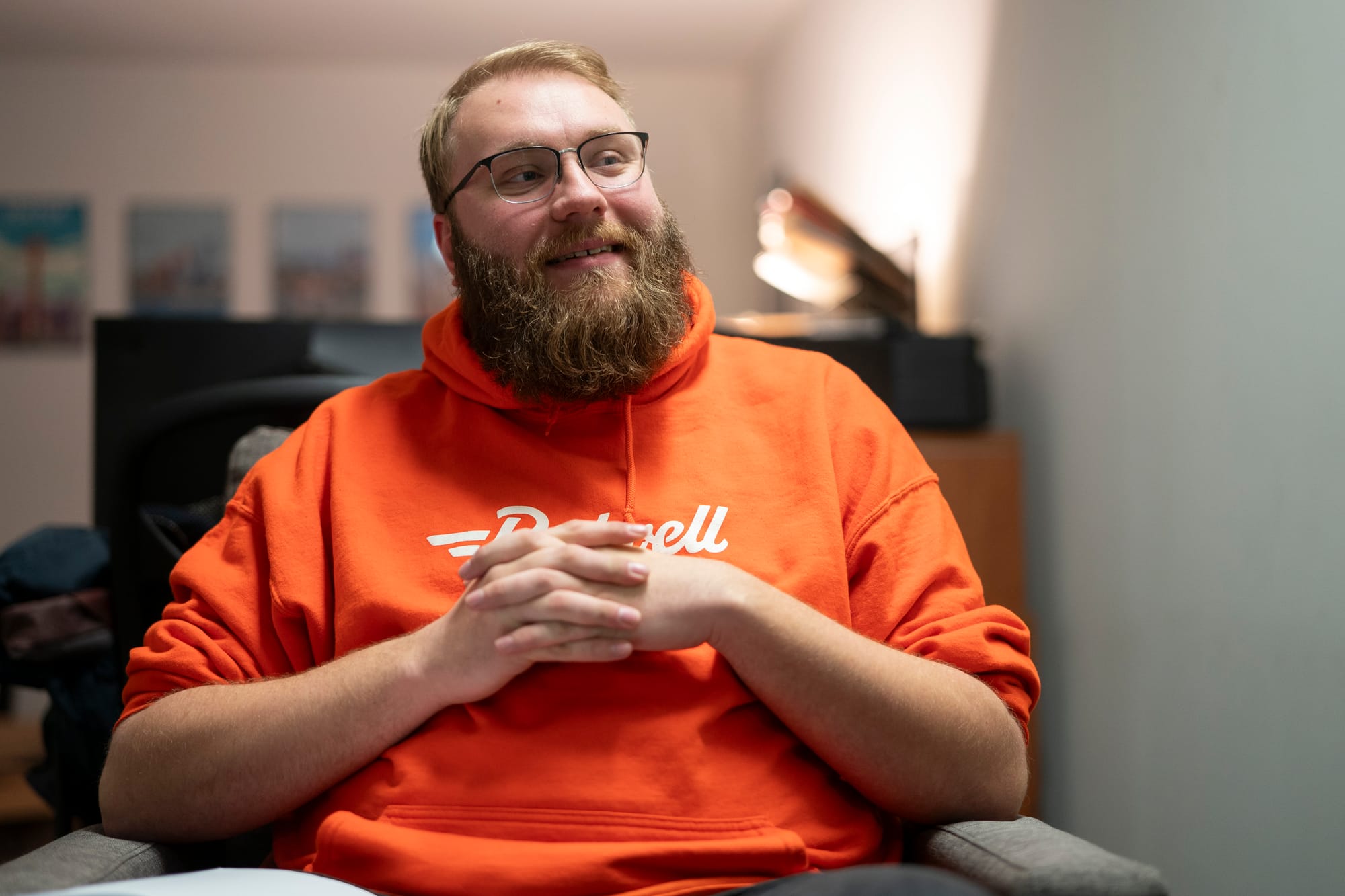 Person with a beard and glasses sits in a chair with hands folded. 