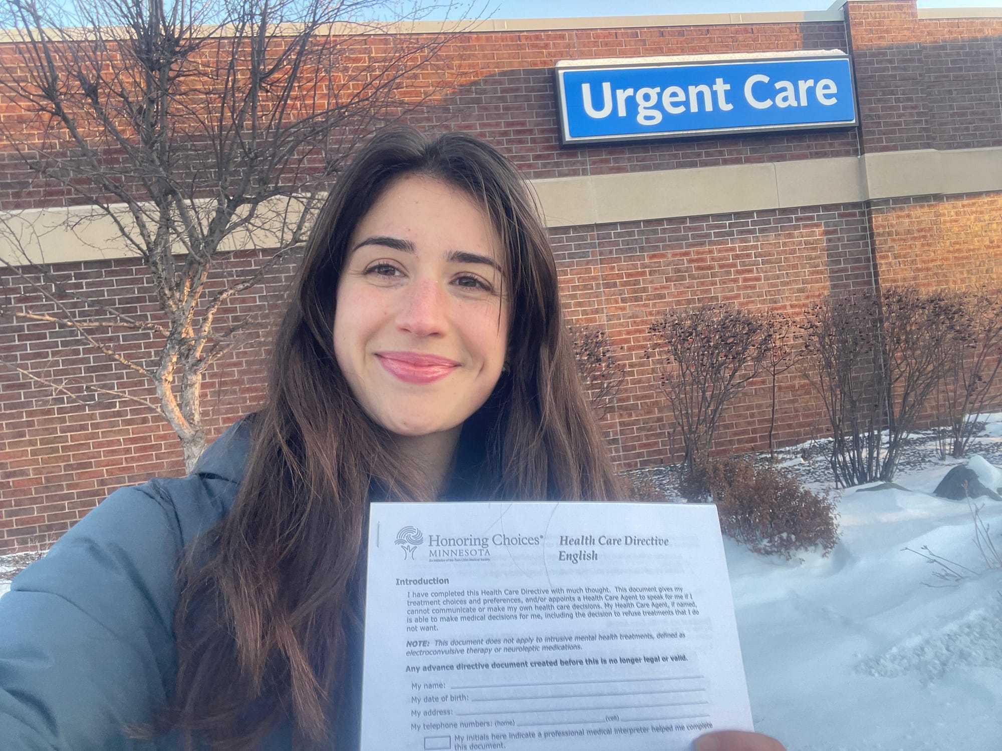 Woman with dark hair holds up advance directive paperwork outside of a health clinic in winter - snow is on the ground. 