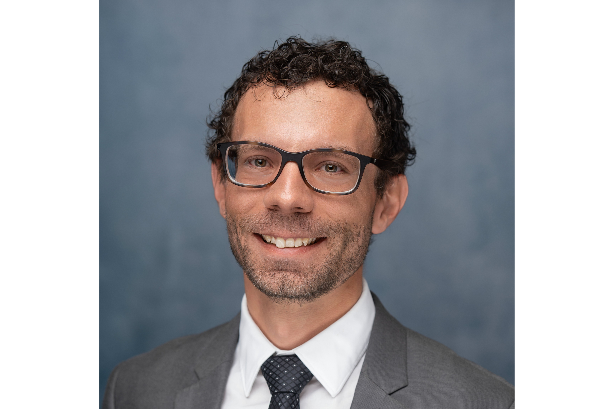 A man with curly dark hair and glasses smiles for a professional portrait. 