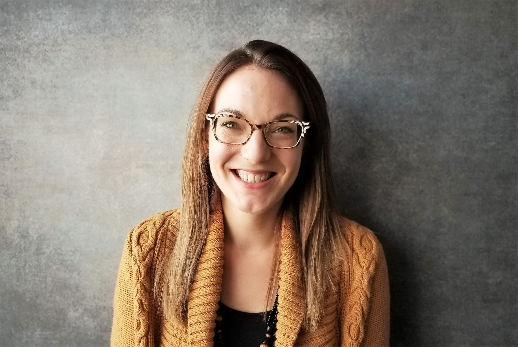 Woman smiles for the camera for a professional headshot. She wears a yellowish-orange sweater over a black shirt. 