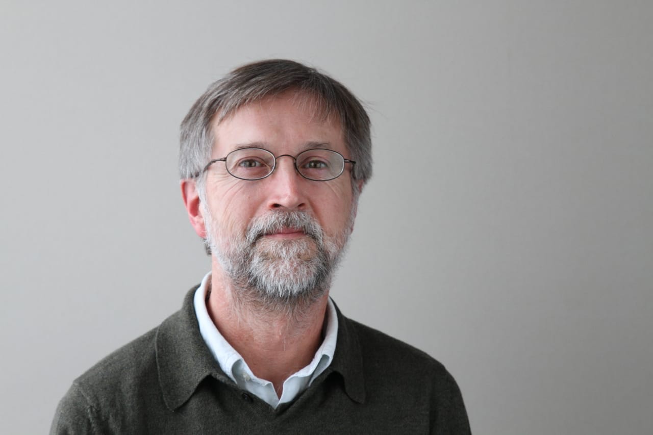 Man smiles for the camera in a professional headshot. He wears a sweater over a button-down shirt. 