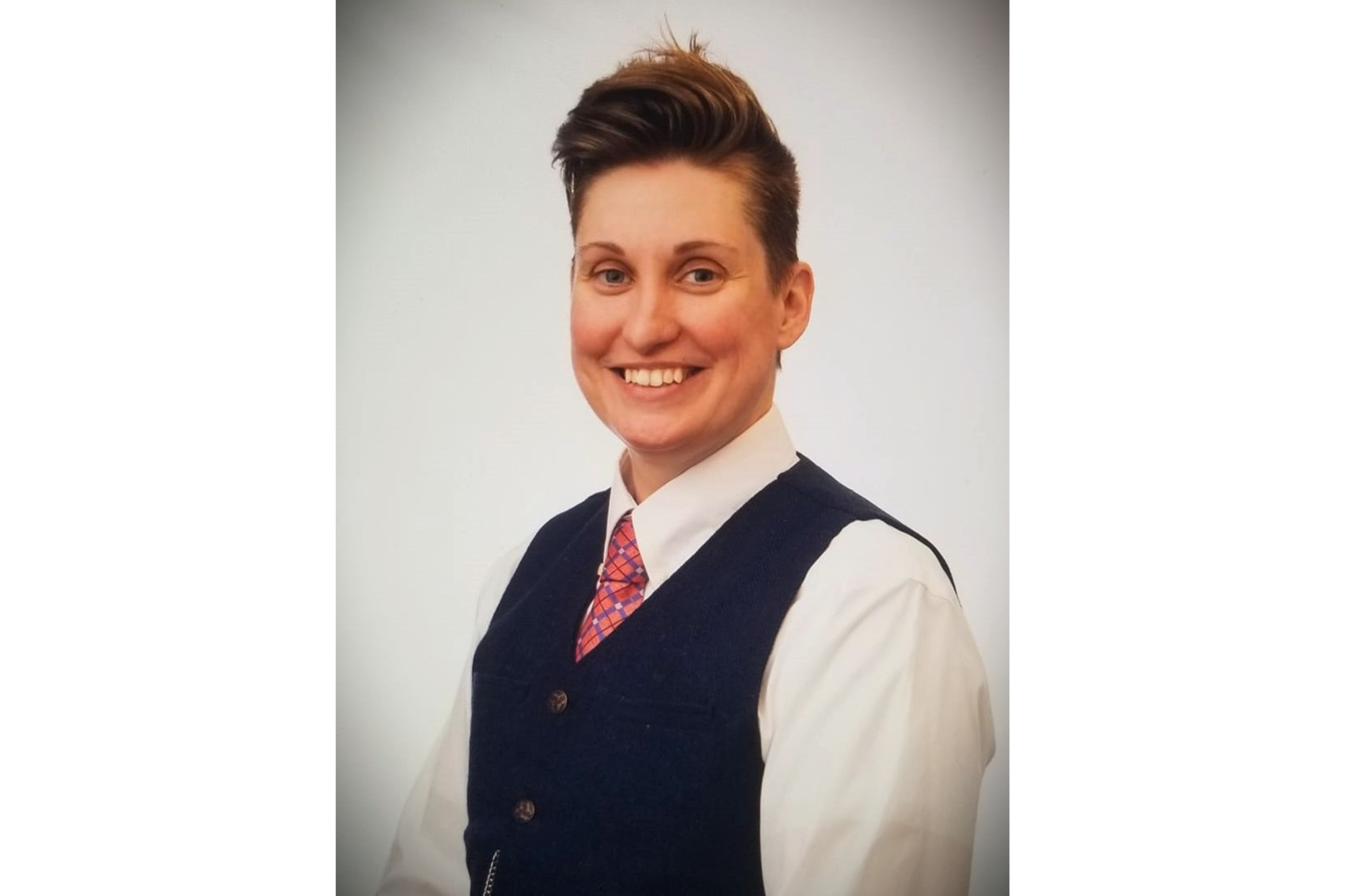 A person poses for a photo wearing a white button down shirt, tie and vest. They smile for the camera. 