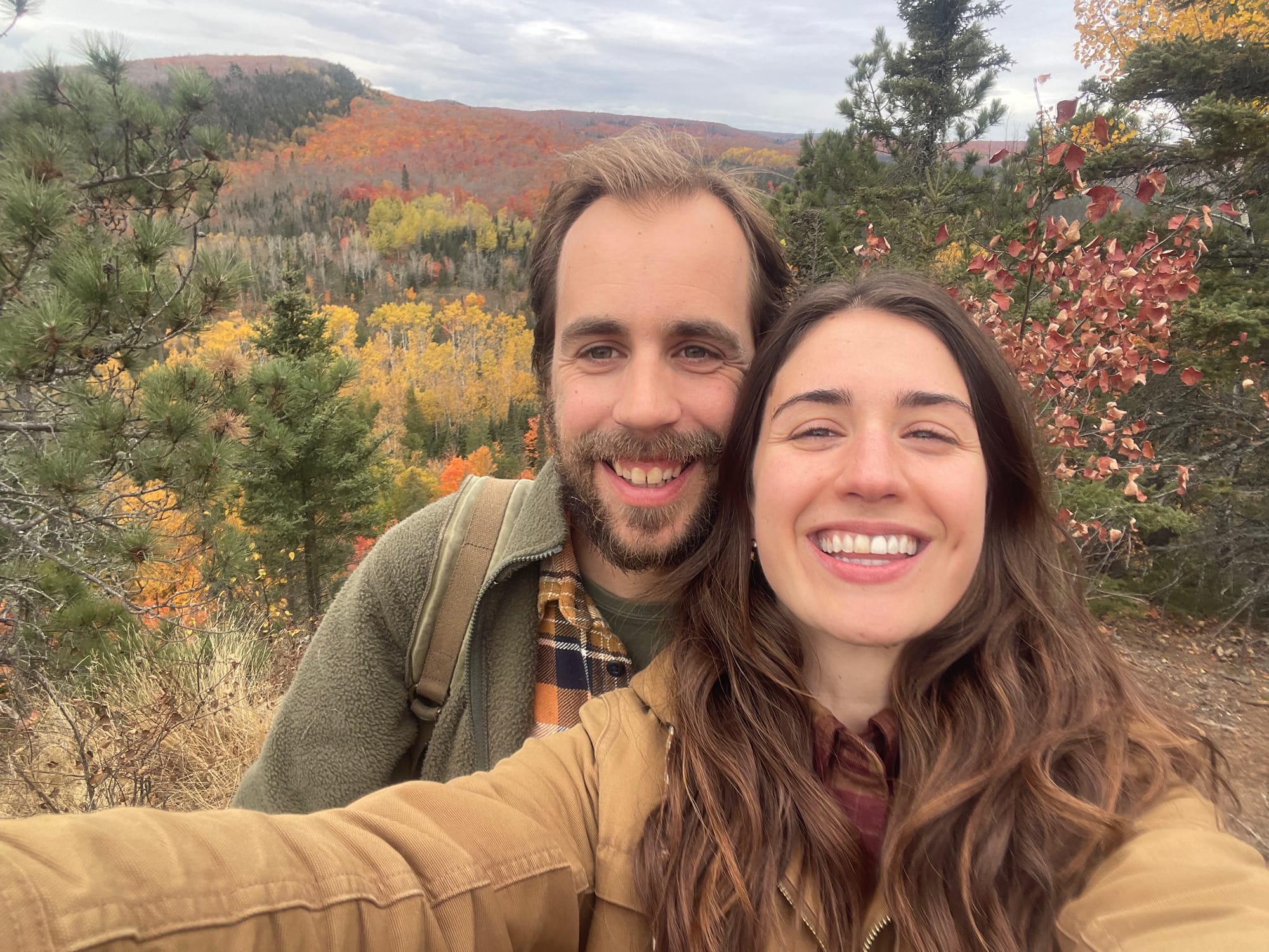 A man and a woman smile for a selfie. The background of the photo has leaves changing color in fall. 