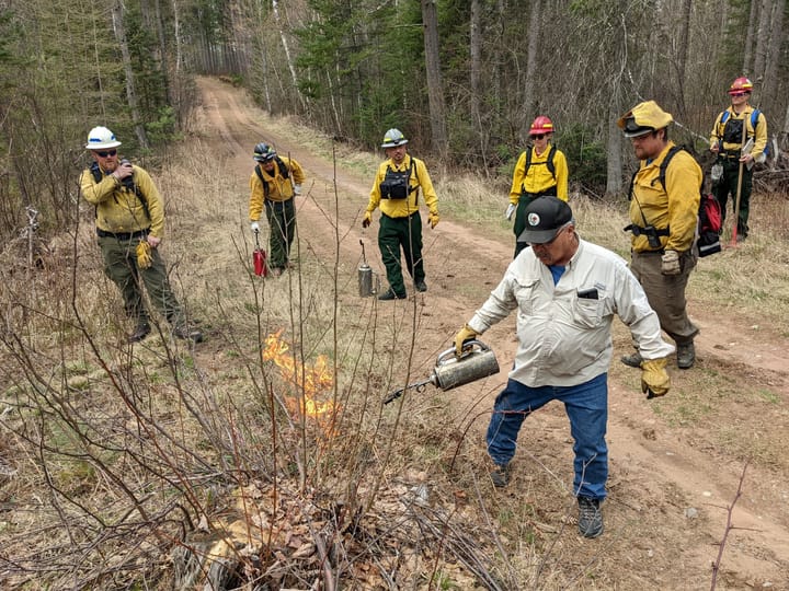 Man starts fire for prescribed burn while firefighters watch. 