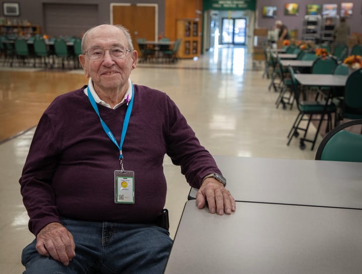 Older man sits at a table while he smiles for a photo. 