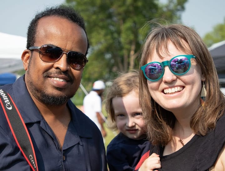 Two adults pose for a photo. A child looks over one person's shoulder. 