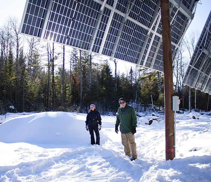 A woman and a man stand on the snowy ground next to a tall solar panel array. Evergreen trees are seen behind them. 