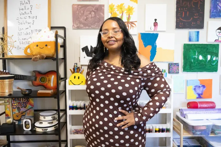 A Somali woman stands in her office. She wears a brown dress with white polka dots. Her long, curly hair falls around her shoulders. She wears clear-rimmed glasses and smiles.