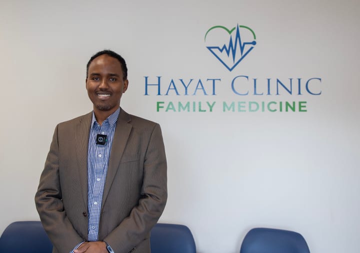 A man smiles for a photo inside his medical clinic. He wears a blue striped button-down shirt underneath a gray suit jacket.